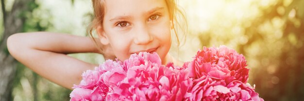 Portret van een gelukkig schattig klein Kaukasisch meisje van zeven jaar, houdt een boeket roze pioenrozen in volle bloei in handen en trekt haar haar recht op de achtergrond van de natuur. spandoek. gloed