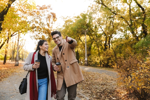 Portret van een gelukkig stel man en vrouw van 20 jaar die afhaalkoffie drinken uit papieren bekers tijdens het wandelen in het herfstpark