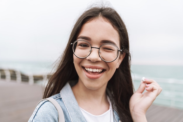 Portret van een gelukkig tienermeisje met een bril die lacht en knipoogt terwijl ze langs de houten pier aan de kust loopt