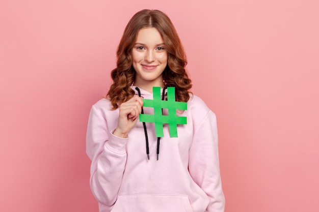 Portret van een gelukkig tienermeisje met krullend haar in hoodie met hashtag-symbool, het promoten van viraal onderwerp in sociaal netwerk, blogtrends taggen. Indoor studio-opname geïsoleerd op roze background