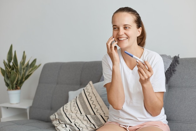 Portret van een gelukkige aantrekkelijke vrouw met een vlecht die een zwangerschapstest in handen houdt, een telefoongesprek voert met de man en hem vertelt dat ze binnenkort ouders zullen zijn, zittend op bed.