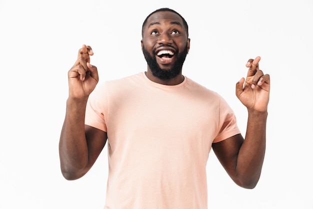 Portret van een gelukkige Afrikaanse man met een t-shirt dat geïsoleerd over een witte muur staat, met de vingers gekruist voor geluk