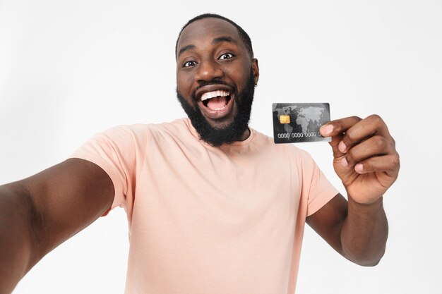 Portret van een gelukkige Afrikaanse man met een t-shirt dat geïsoleerd over een witte muur staat, selfie neemt, creditcard toont