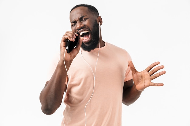 Portret van een gelukkige Afrikaanse man met een t-shirt die geïsoleerd over een witte muur staat, naar muziek luistert met een koptelefoon, een mobiele telefoon vasthoudt