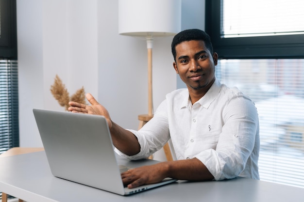 Portret van een gelukkige Afrikaanse mannelijke ondernemer die een laptop gebruikt en vriendelijk naar de camera kijkt terwijl hij geniet van zijn werk aan een bureau in een moderne kantoorruimte