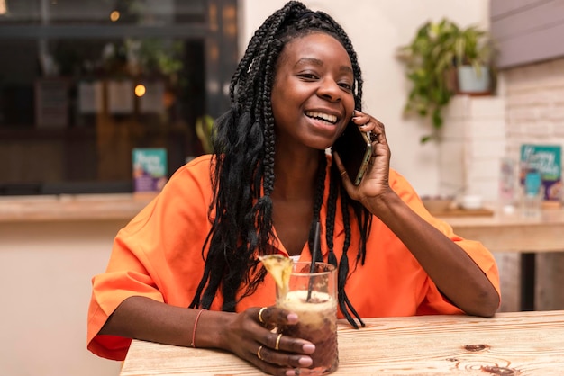 portret van een gelukkige afro-amerikaanse vrouw die een drankje vasthoudt en telefonisch praat in een bar