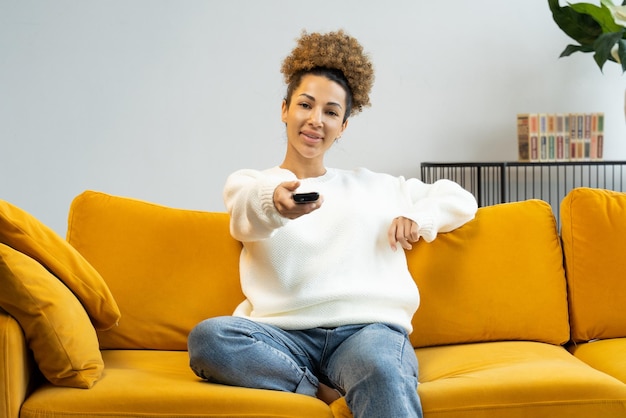 Portret van een gelukkige Afro-Amerikaanse vrouw die op de bank zit en van zender wisselt op de tv