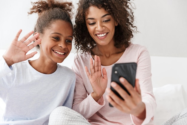 Portret van een gelukkige Afro-Amerikaanse vrouw en haar dochtertje die mobiel samen op de bank thuis gebruiken