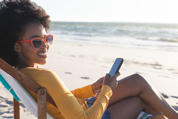 Portret van een gelukkige Afro-Amerikaanse vrouw in een zonnebril die smartphone gebruikt op zonnig strand. Zomer, ontspanning, communicatie, vrije tijd en vakantie, onveranderd.