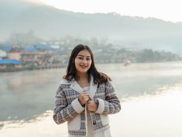 Portret van een gelukkige Aziatische vrouw in een winterjas met mist die 's ochtends opkomt op het uitzicht op het meer in het dorp Ban Rak Thai, Mae Hong Son in Thailand.