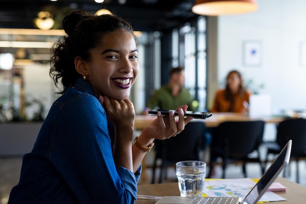 Foto portret van een gelukkige biracial casual zakenvrouw met een laptop die op een smartphone praat in de kantoorlounge