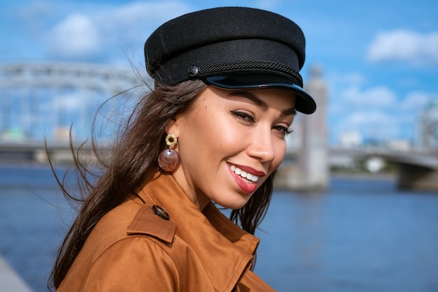 Portret van een gelukkige blanke vrouw aan de kade van de rivier in een zwarte pet en een bruin jasje op een zonnige dag
