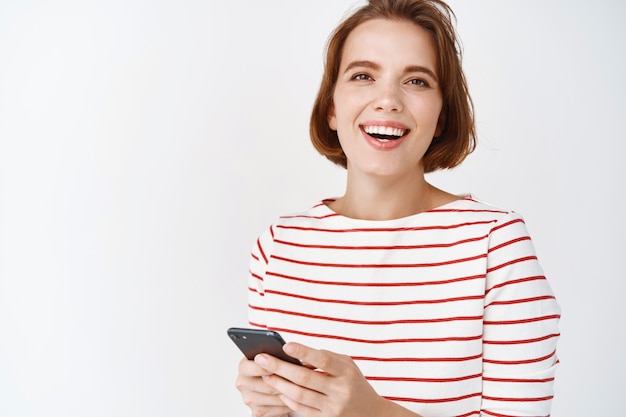 Portret van een gelukkige blanke vrouw in gestreepte blouse, lachen en glimlachen, chatten op smartphone en glimlachen, staande tegen een witte muur