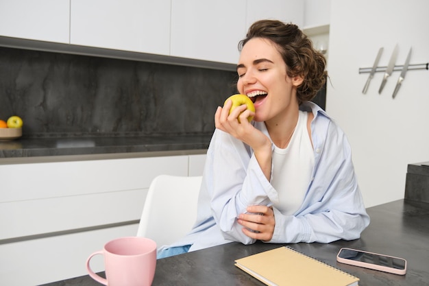 Portret van een gelukkige brunette vrouw die een appel bijt en een gezonde snack eet terwijl ze in de keuken zit Mensen en gezond dieetconcept