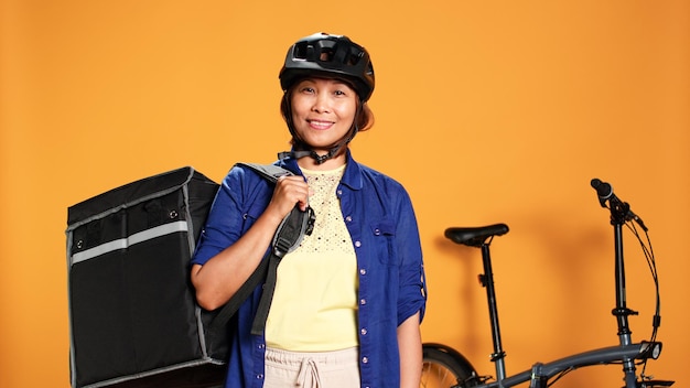 Portret van een gelukkige glimlachende Aziatische fietser met een thermische rugzak geïsoleerd over een oranje achtergrond van een studio Vriendelijke koerier die takeaway eten aan de klant levert