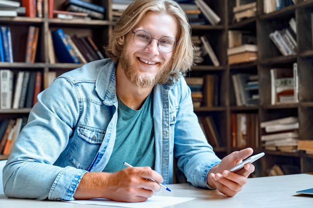 Portret van een gelukkige glimlachende jonge student aan de bibliotheekbalie met een moderne smartphone