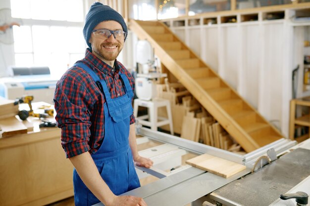 Foto portret van een gelukkige glimlachende timmerman in overalls en beschermende bril die op de werkplek staat en klaar is om op de cirkelzaagwerkbank te werken