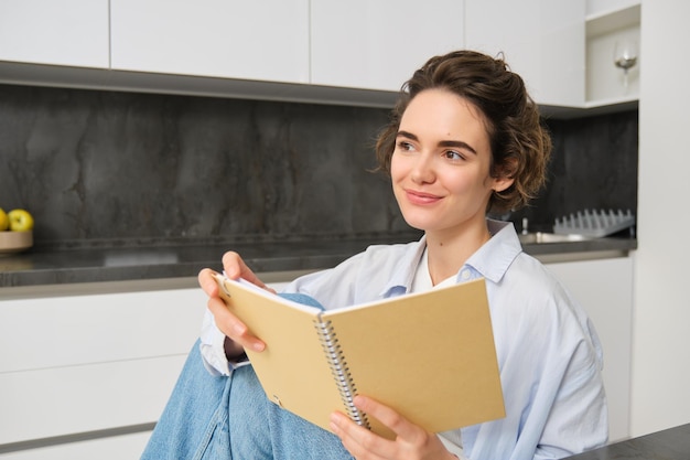 Portret van een gelukkige glimlachende vrouw die een notitieboekje vasthoudt en in haar keukenlezingsdagboek studeert