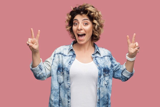 Portret van een gelukkige grappige jonge vrouw met krullend haar in een casual shirt dat met een gebaar van de overwinning of een vredesteken staat en naar de camera kijkt met een verbaasd gezicht. indoor studio opname, geïsoleerd op roze achtergrond.