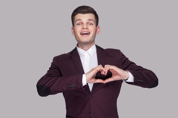 Portret van een gelukkige grappige knappe jongeman in violet pak en wit overhemd, staand, kijkend naar camera met hartgebaar teken. indoor studio opname, geïsoleerd op een grijze achtergrond.