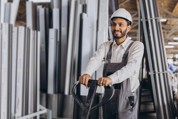 Portret van een gelukkige hindoe-arbeider in een witte helm en overall met een hydraulische vrachtwagen tegen een achtergrond van een fabriek en aluminium frames