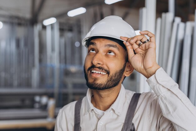 Portret van een gelukkige hindoe-arbeider in een witte helm en overall met een hydraulische vrachtwagen tegen een achtergrond van een fabriek en aluminium frames