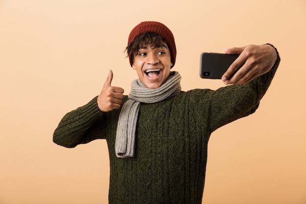 Portret van een gelukkige jonge Afrikaanse man gekleed in herfstkleren geïsoleerd over beige muur, een selfie nemen, duimschroef opwaarts gebaar