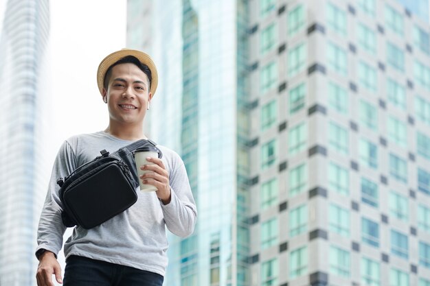 Portret van een gelukkige jonge aziatische man in een strohoed met een afhaalkoffiekop die tegen hoge gebouwen in de grote stad staat