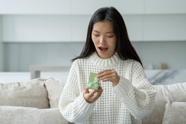 Portret van een gelukkige jonge aziatische vrouw die een geschenkdoos opent terwijl ze op de bank zit in haar woonkamer