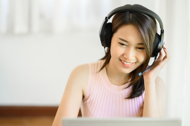 Portret van een gelukkige jonge Aziatische vrouw die een headset draagt terwijl ze thuis aan een notebook werkt