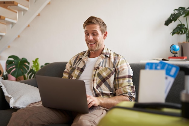 Portret van een gelukkige jonge man die een vakantie voor twee boeken heeft vliegtickets kijkt naar een laptop zit in de woonkamer
