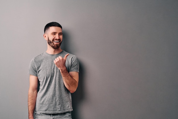 Portret van een gelukkige jonge man in grijze t-shirt wijzende vingers weg naar kopieerruimte geïsoleerd over grijze achtergrond