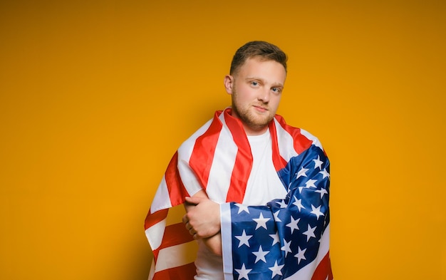 Portret van een gelukkige jonge man met een baard in vrijetijdskleding die de Amerikaanse vlag op een geel houdt