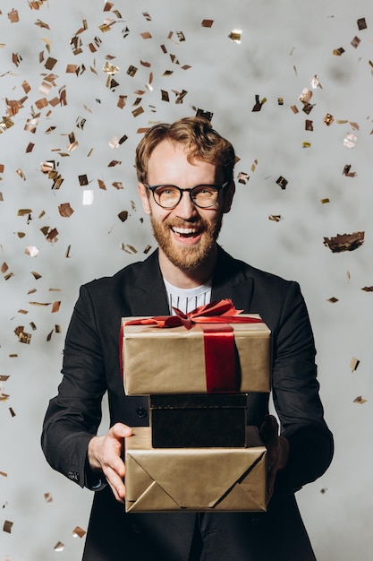 Portret van een gelukkige jonge man met een grote glimlach die geschenken vasthoudt terwijl gouden confetti vallenxA