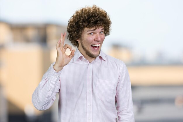 Foto portret van een gelukkige jonge man met krullend haar toont goed gebaar teken wazig stedelijke achtergrond