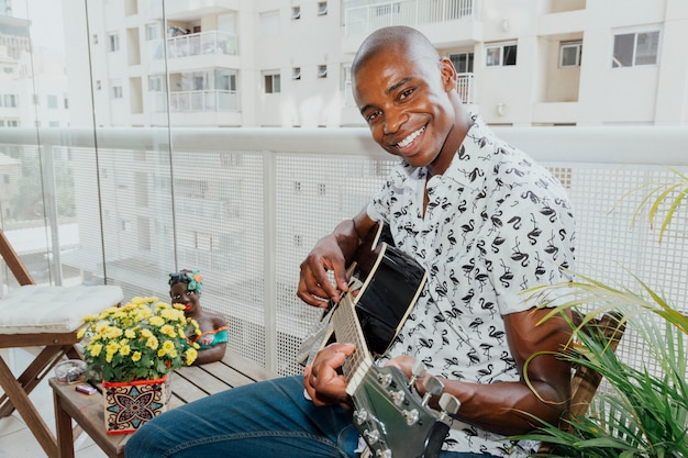 Portret van een gelukkige jonge man zit op het balkon spelen gitaar kijken camera