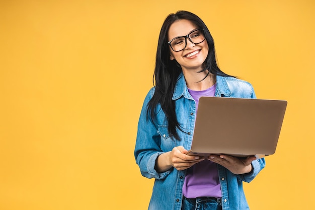 Portret van een gelukkige jonge, mooie verraste vrouw met een bril die met een laptop staat geïsoleerd op een gele achtergrond Ruimte voor tekst