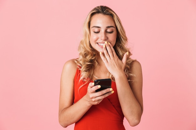 Portret van een gelukkige jonge opgewonden schattige blonde vrouw poseren geïsoleerd over roze muur met behulp van mobiele telefoon