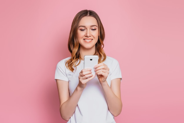 Portret van een gelukkige jonge сaukasische vrouw in een wit T-shirt met behulp van mobiele telefoon geïsoleerd over roze muur studente met golvend krullend haar kijkt naar smartphone en leest een bericht