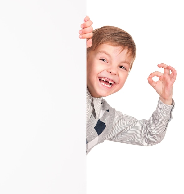 Portret van een gelukkige kleine jongen met een leeg bord geïsoleerd op een witte achtergrond Glimlachend kind met een banier en een ok gebaar Grappig kind dat achter een leeg paneel of bord kijkt