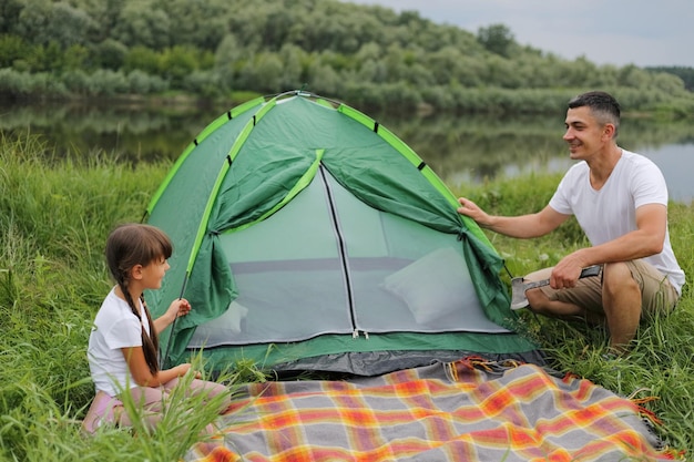 Portret van een gelukkige knappe man met casual kleding die zich voordeed in de buurt van een groene tent met zijn schattige dochterfamilie die samen kampeert en geniet van de prachtige natuur