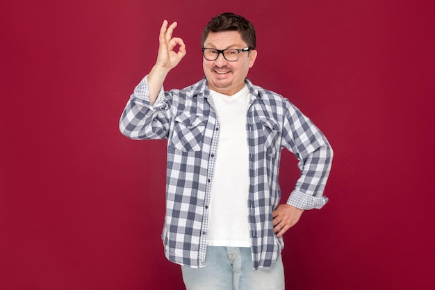 Foto portret van een gelukkige knappe zakenman van middelbare leeftijd in een casual geruit overhemd, een bril met een ok-teken en een brede glimlach die naar de camera kijkt. indoor studio opname, geïsoleerd op donker rode achtergrond.