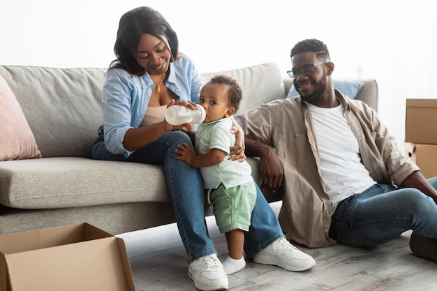 Portret van een gelukkige liefhebbende Afro-Amerikaanse vrouw die haar kind voedt met een babyfles, schattige zwarte peuter drinkwater of melk. Ouders zitten op de bank pakketjes uit te pakken op verhuisdag