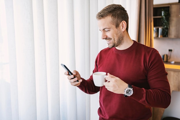 Portret van een gelukkige man die thuis koffie drinkt en telefoon gebruikt