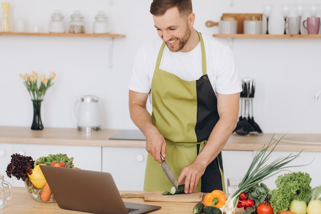 Portret van een gelukkige man thuis, een man die groentesalade kookt die naar de camera kijkt en glimlacht terwijl hij groenten snijdt met een laptop voor online kooktraining