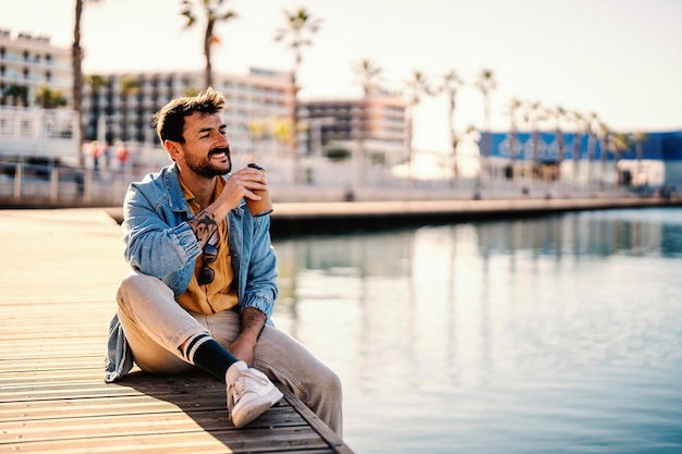 Portret van een gelukkige man zittend op de pier naast het water en genietend van zijn koffie