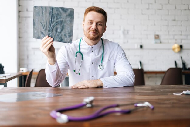 Portret van een gelukkige mannelijke arts in een wit uniform die de computertomografie van de hersenen onderzoekt aan een bureau in het ziekenhuiskantoor