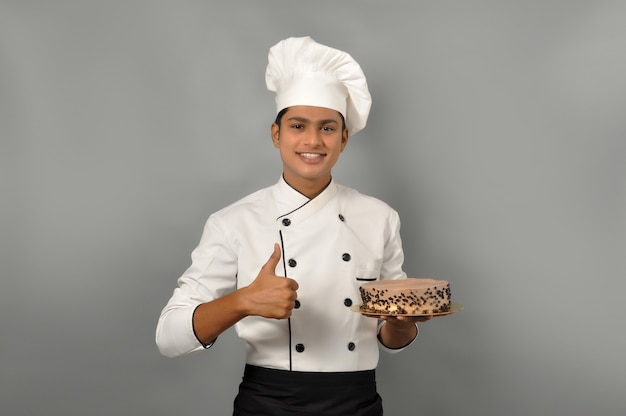 Portret van een gelukkige mannelijke chef-kok gekleed in uniforme holdingsplaat met chocoladetaart met duimen omhoog