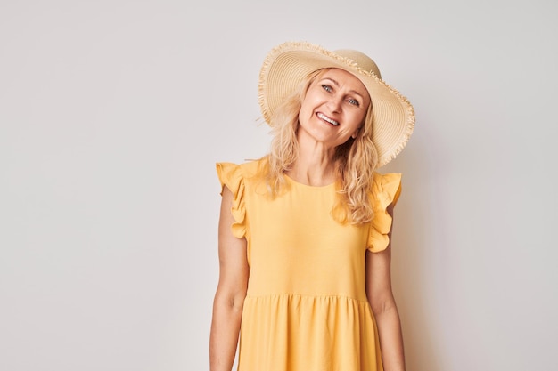 Portret van een gelukkige mooie blonde volwassen vrouw in een gele zomerjurk en een strohoed die vrolijk glimlacht geïsoleerd op een witte achtergrond van een studio Vakantie- en reisconcept