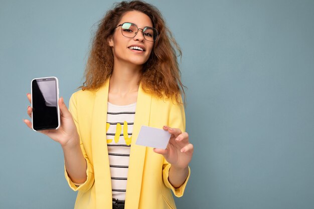 Portret van een gelukkige mooie vrouw met een gele jas en een optische bril die op een achtergrondmuur is geïsoleerd en een telefoon toont met een leeg scherm en een creditcard die naar de camera kijkt